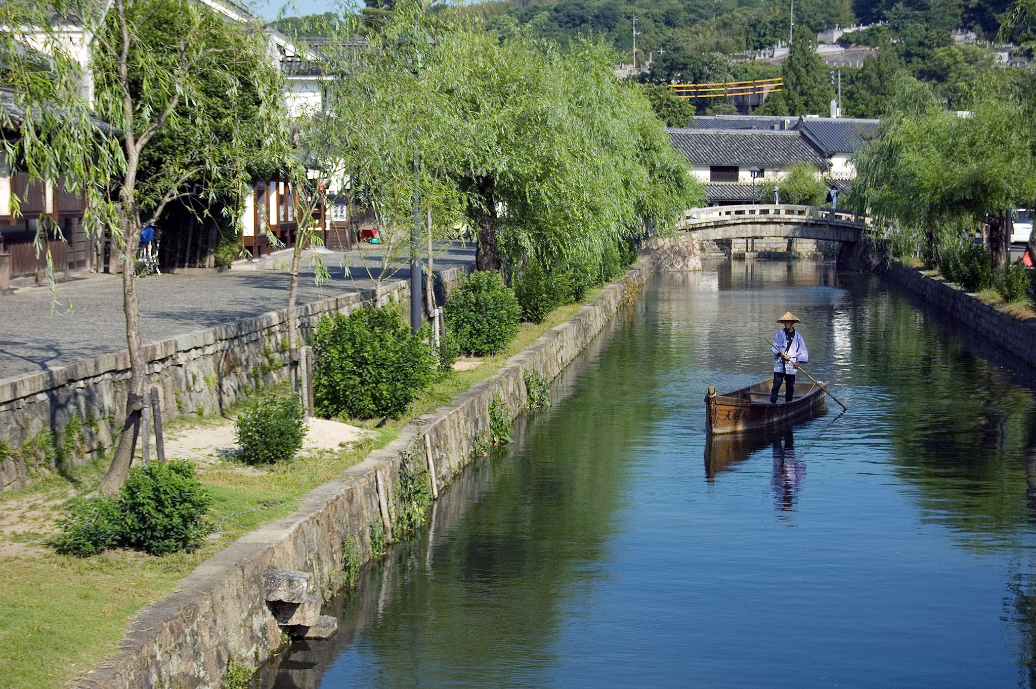 In the idyllic "Kurashiki Bikan Historical Quarter," a man peacefully rows a boat along a serene canal in a small town.