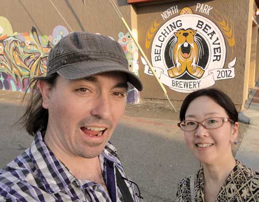 A couple stands before a brewery mural, posing for a photo. The man and women do Japanese & English beer marketing.